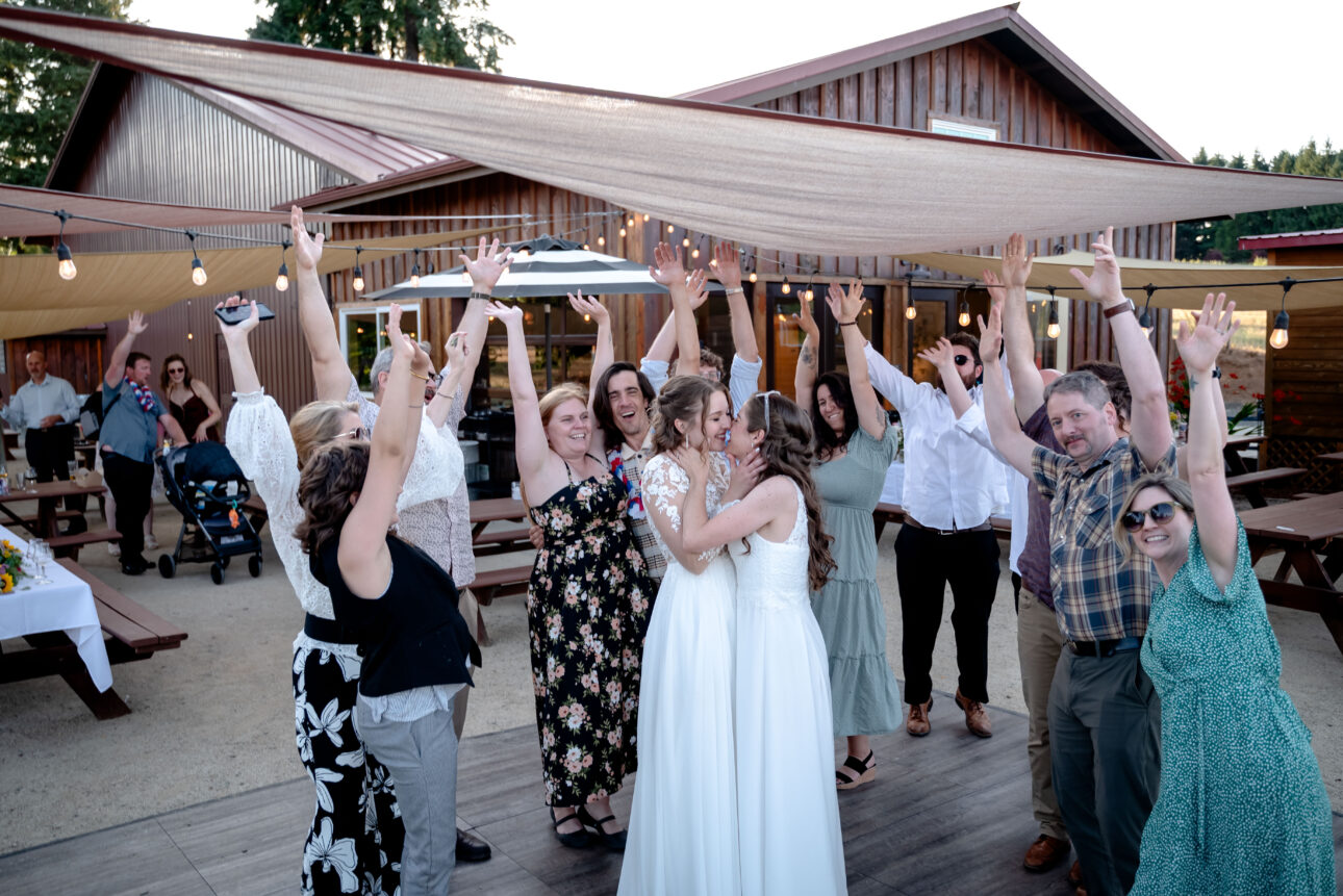 Two brides kiss on a wedding dance floor surrounded by friends and family celebrating their love story.
